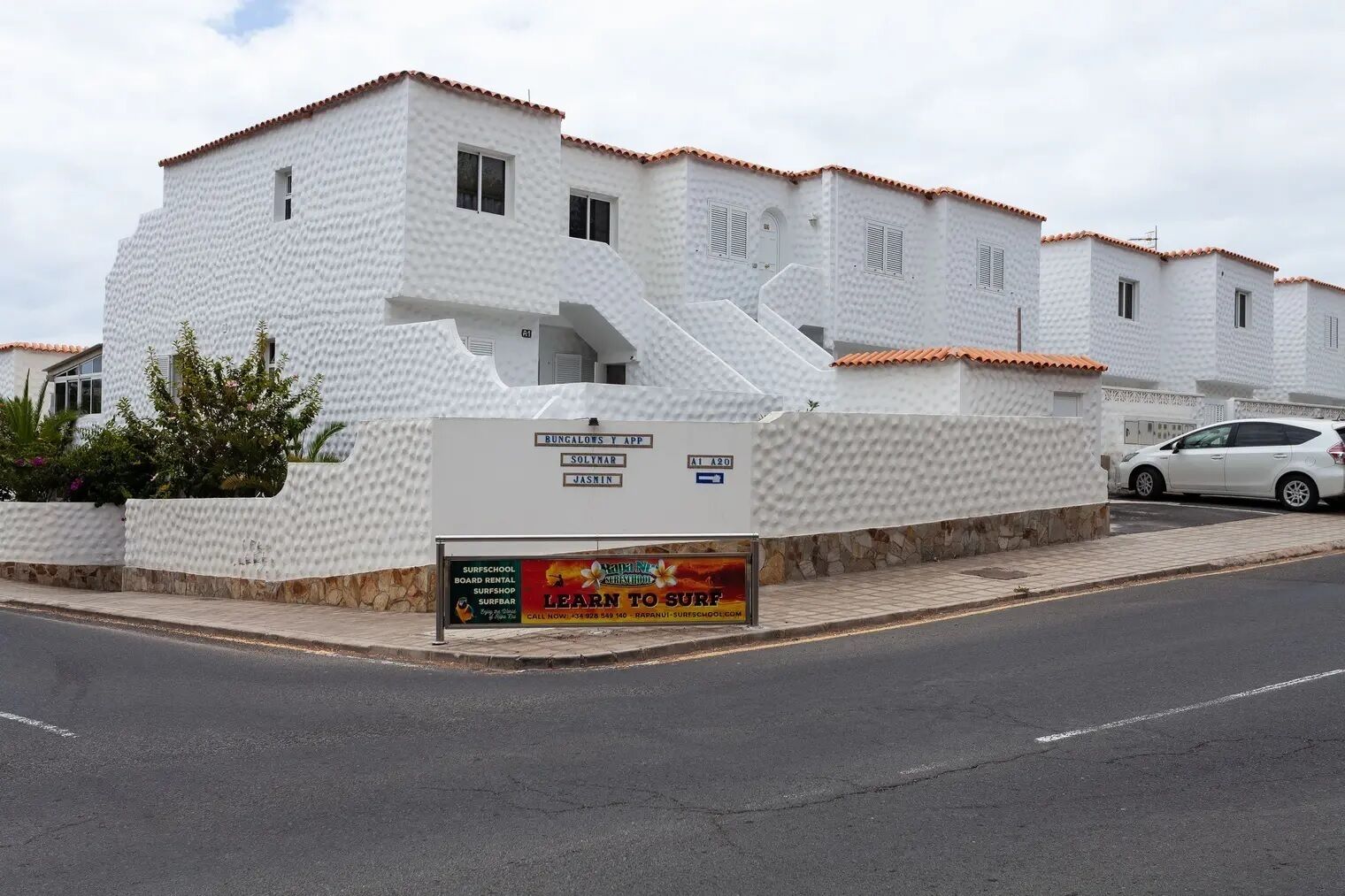 A white building with a tiled roof, a signboard advertising surf school and board rental, and a parked car.