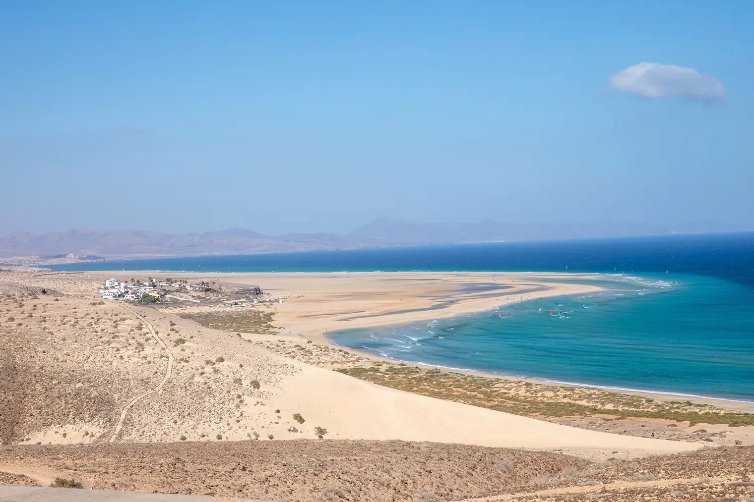 A coastal landscape with a sandy beach, a small settlement, and distant mountains.