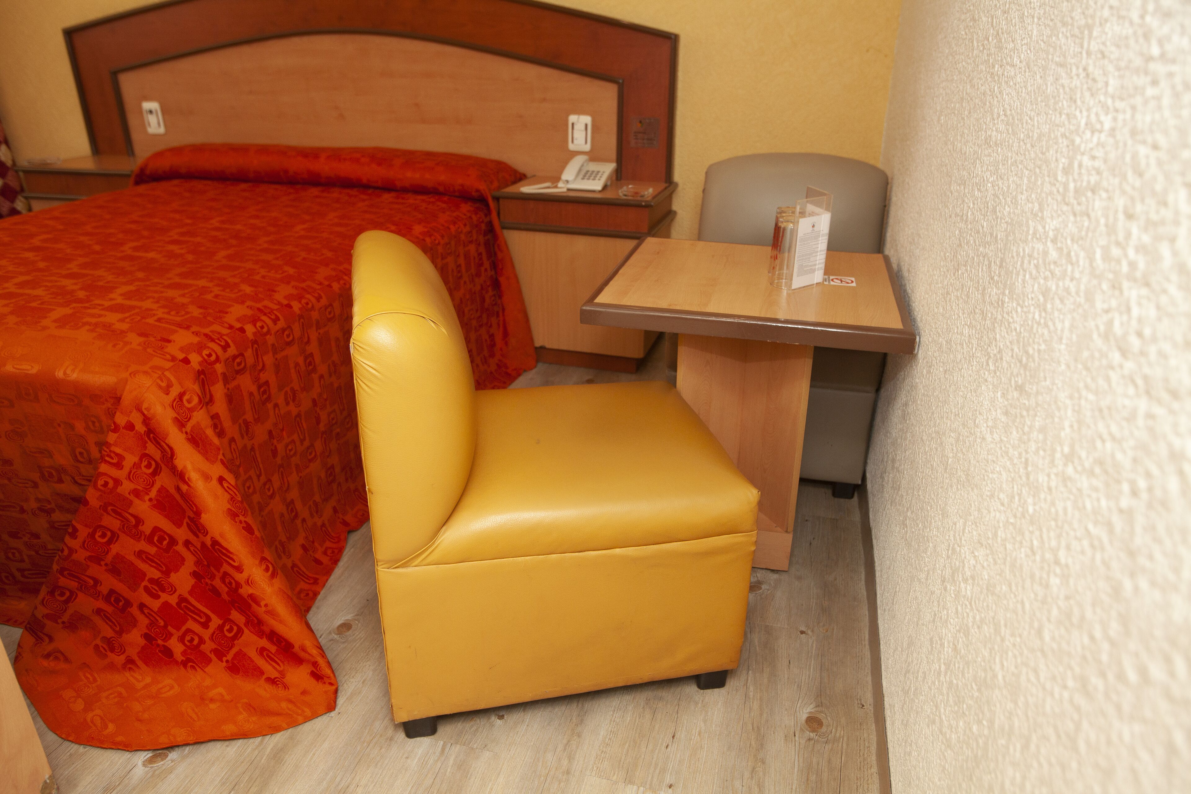 Una habitación de hotel con una cama, una silla amarilla, una mesita pequeña y una mesita de noche con un teléfono.