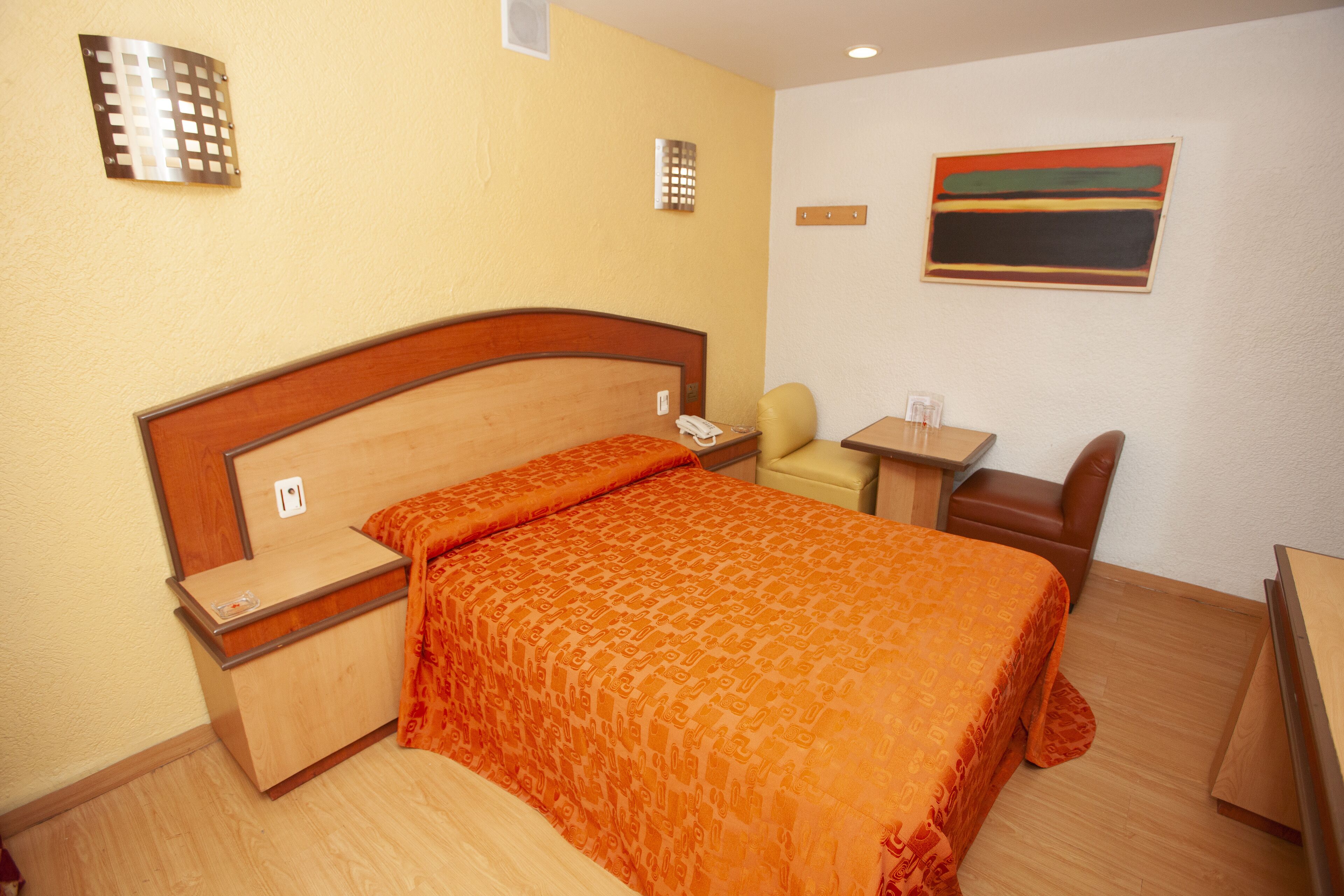 A hotel room with a bed, bedside table, chair, desk, and a painting on the wall.
