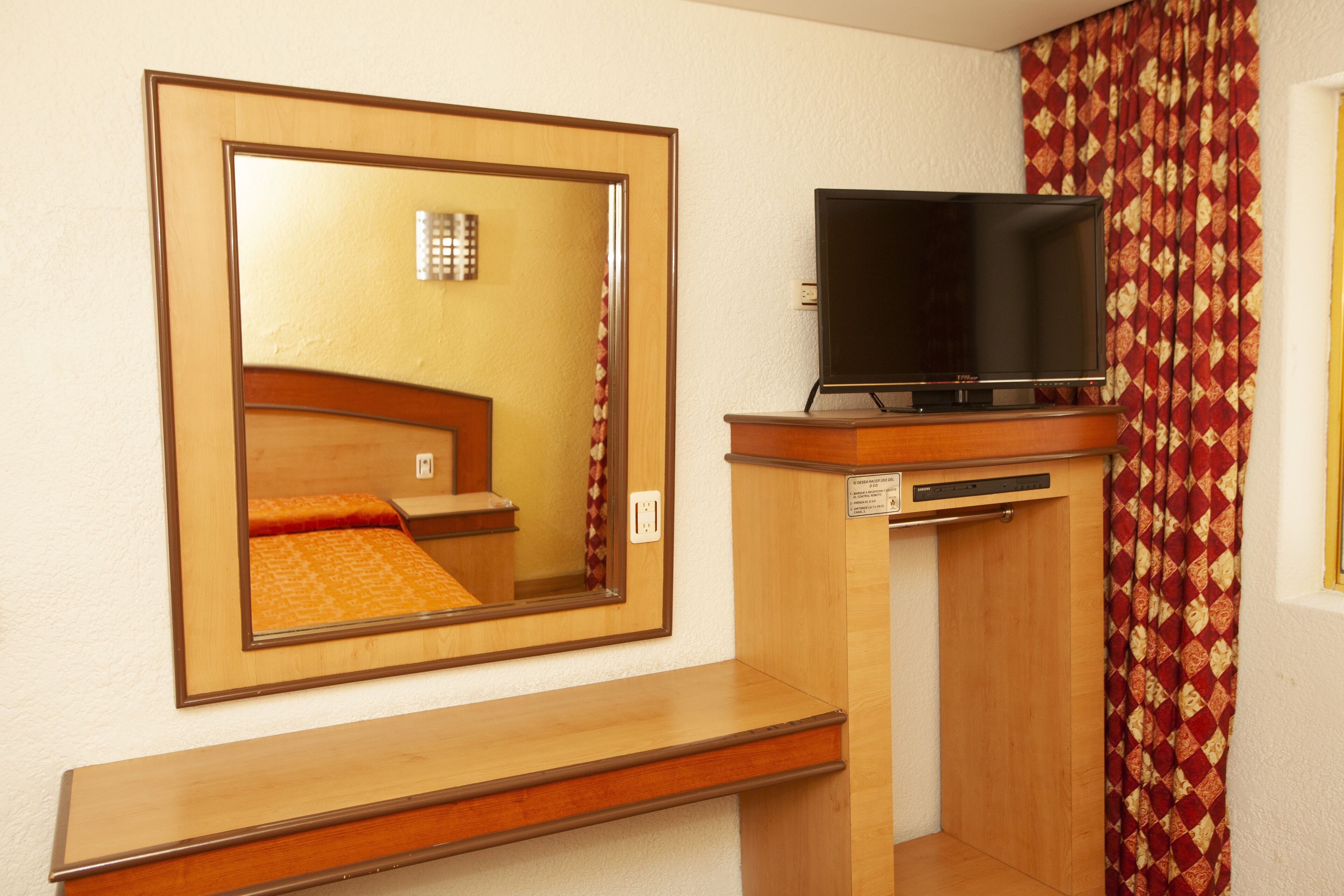A hotel room with a wooden dresser, a flat-screen TV on top, a large mirror, and a patterned curtain.