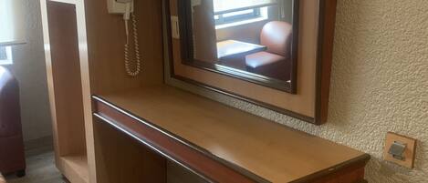 Une pièce de taille modeste, avec un bureau en bois, un téléviseur à écran plat fixé au mur au-dessus, une petite poubelle et une prise électrique sur le mur.