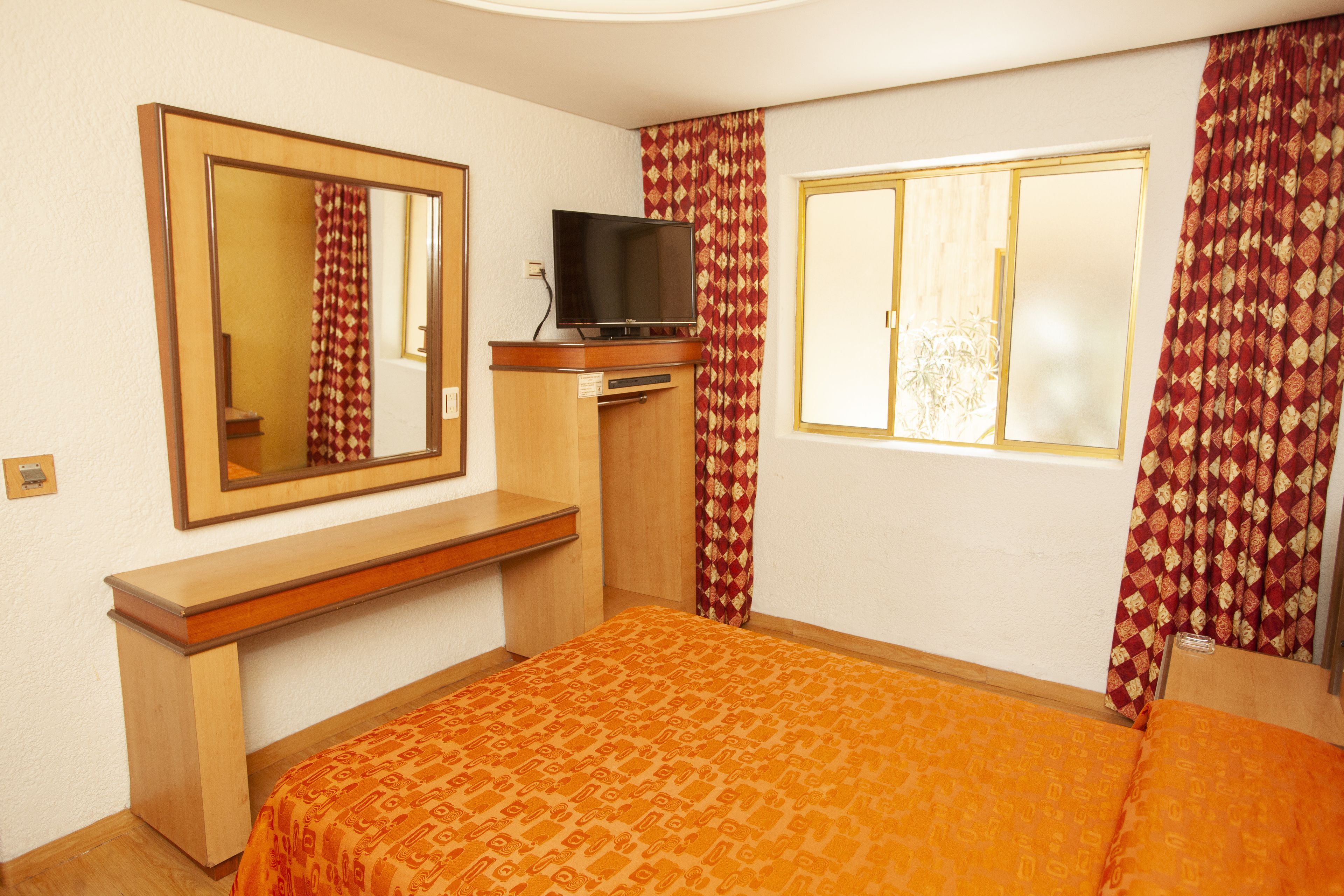 A hotel room with a bed, a TV on a wooden cabinet, a mirror, and patterned curtains.