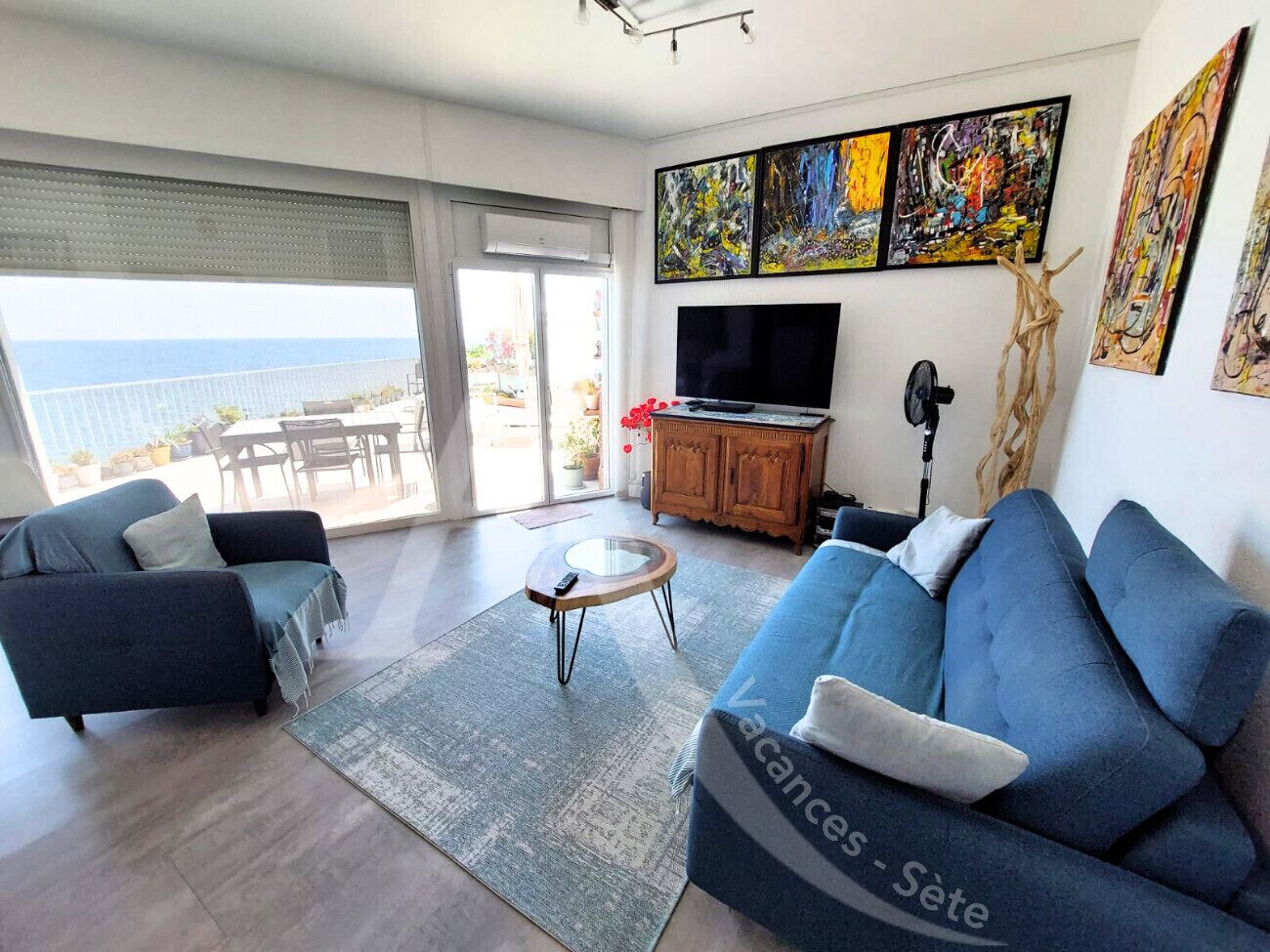 Una sala de estar con un sofá, un sillón y una mesa de centro. Hay un televisor, obras de arte en la pared y vistas al exterior.