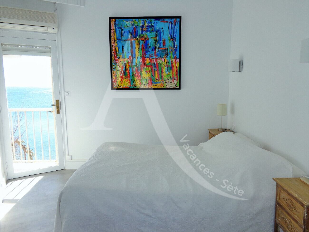 A bedroom with a bed, a nightstand, a wall-mounted painting, and a balcony with a view of the sea.