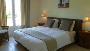 A neatly made bed with a white and beige bedspread, two bedside lamps, a wicker chair, and a door with curtains. - Villa in France with Pool and Private Garden (Vasles)