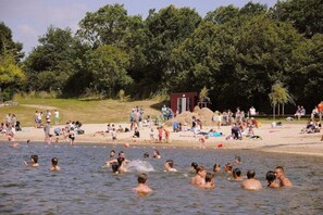 Una spiaggia affollata con persone che nuotano e prendono il sole, una piccola struttura e alberi fitti sullo sfondo.