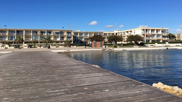 A waterfront hotel with a wooden pier extending into the water, featuring multiple balconies and a pool area.