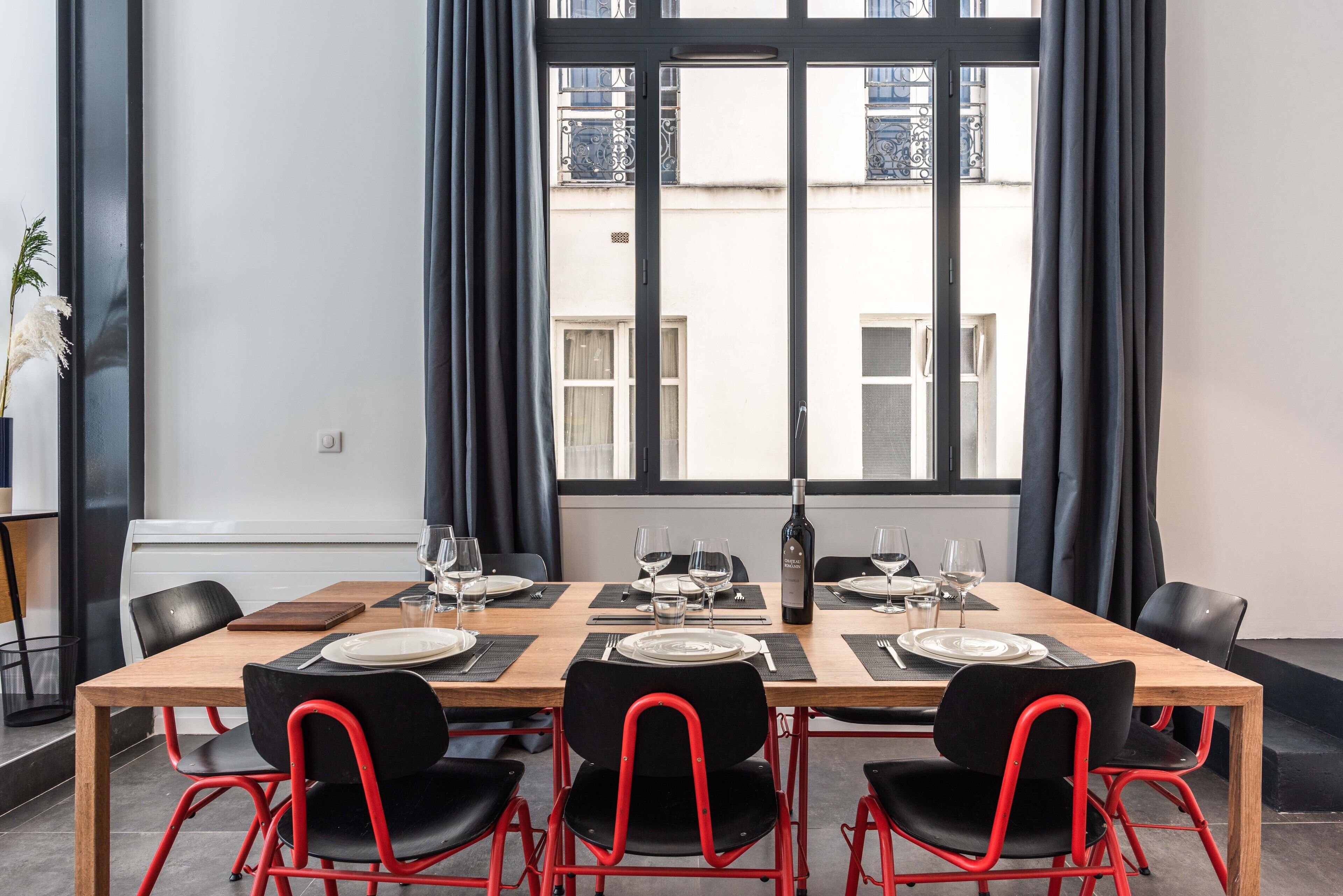 A dining area with a wooden table set for four, black chairs with red frames, and a view of buildings through large windows.