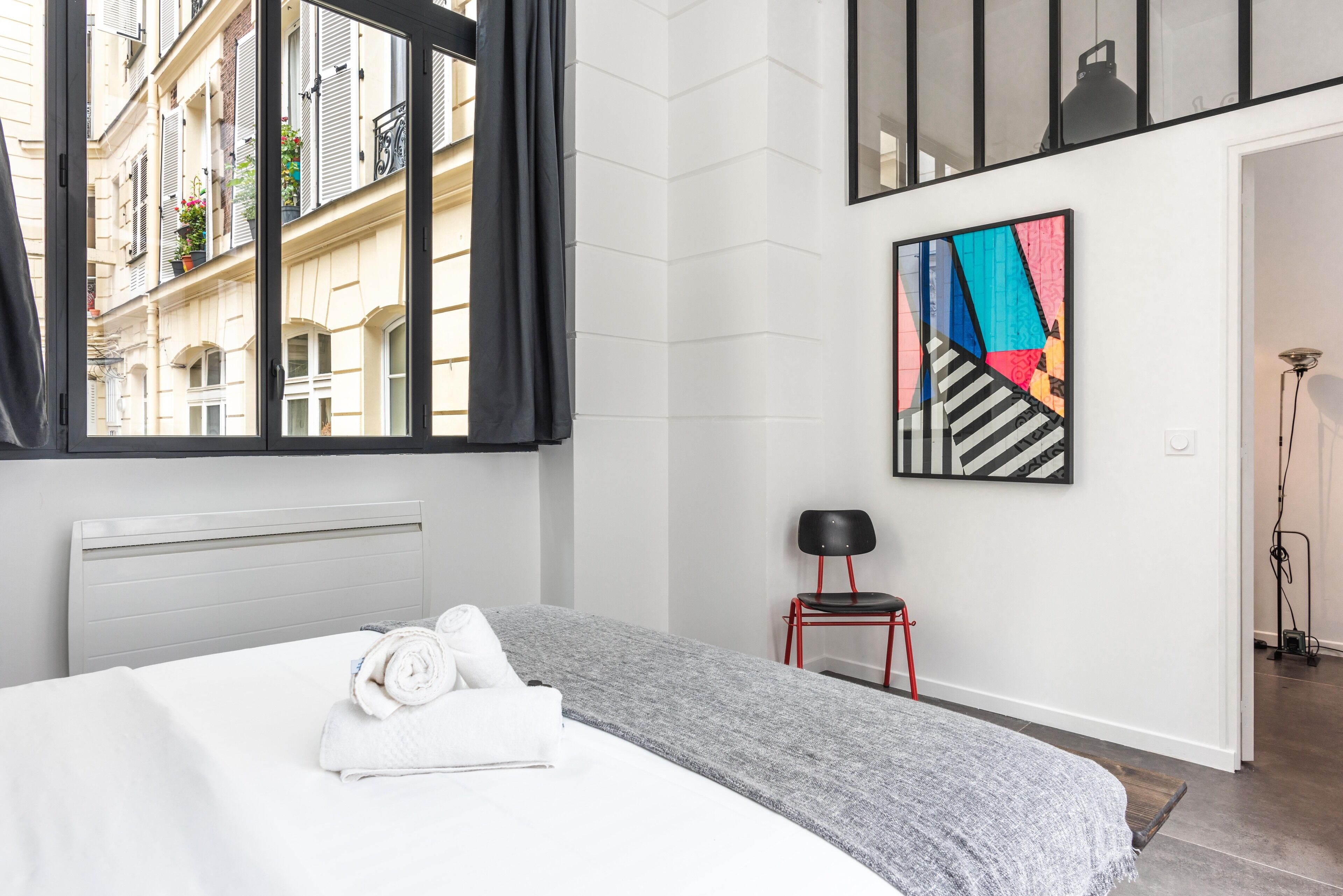 Una habitación de hotel moderna con una cama, un ventanal con cortinas, una silla roja y un cuadro abstracto colorido en la pared.