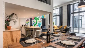 Un espace repas moderne avec une table en bois dressée pour six personnes, une cuisine en arrière-plan et de grandes fenêtres donnant sur la rue.