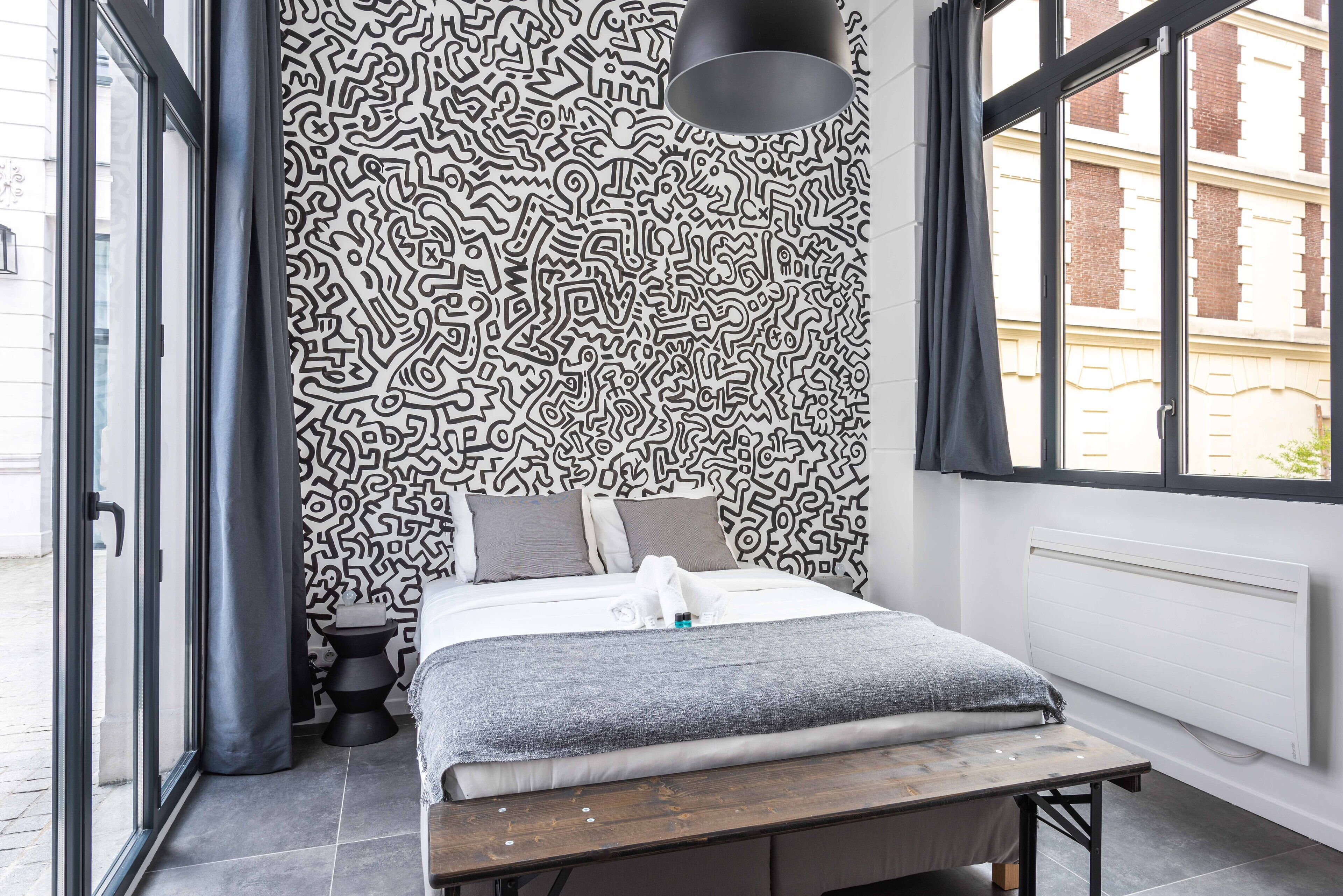 Une chambre moderne avec un mur à motifs, un lit recouvert d’une couverture grise, une table de chevet et une fenêtre avec des rideaux.