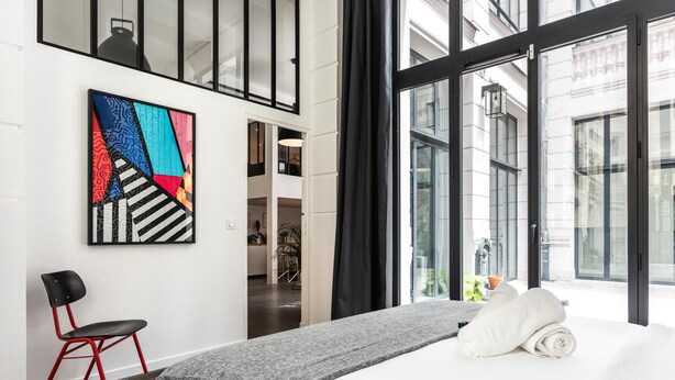 Une chambre d’hôtel moderne avec une grande fenêtre, un lit recouvert de draps blancs, un plaid gris, une chaise rouge et noire, et un tableau abstrait aux couleurs vives accroché au mur.