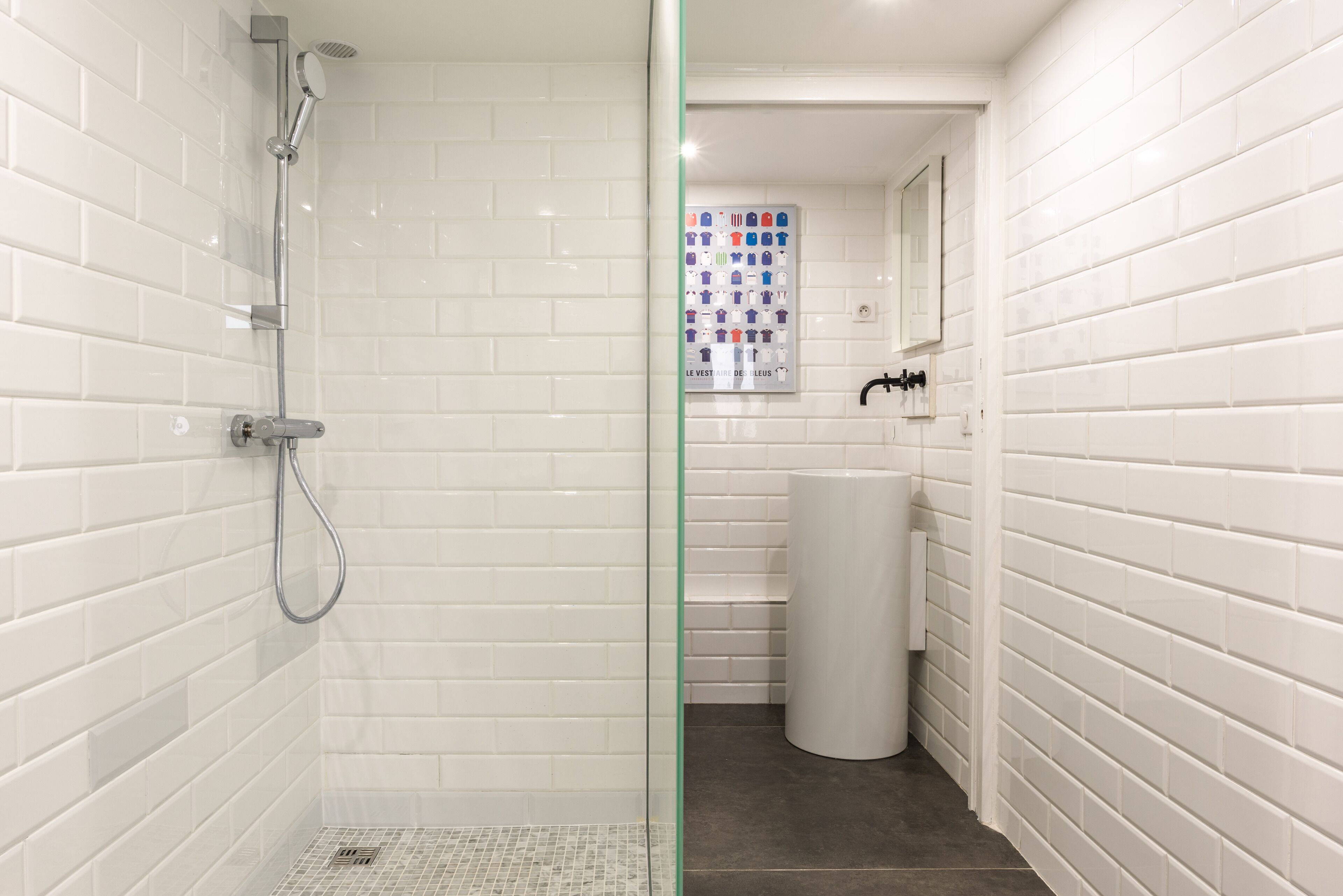 A bathroom with white tiled walls, a shower with a handheld showerhead, a toilet, and a sink with a mirror above it.