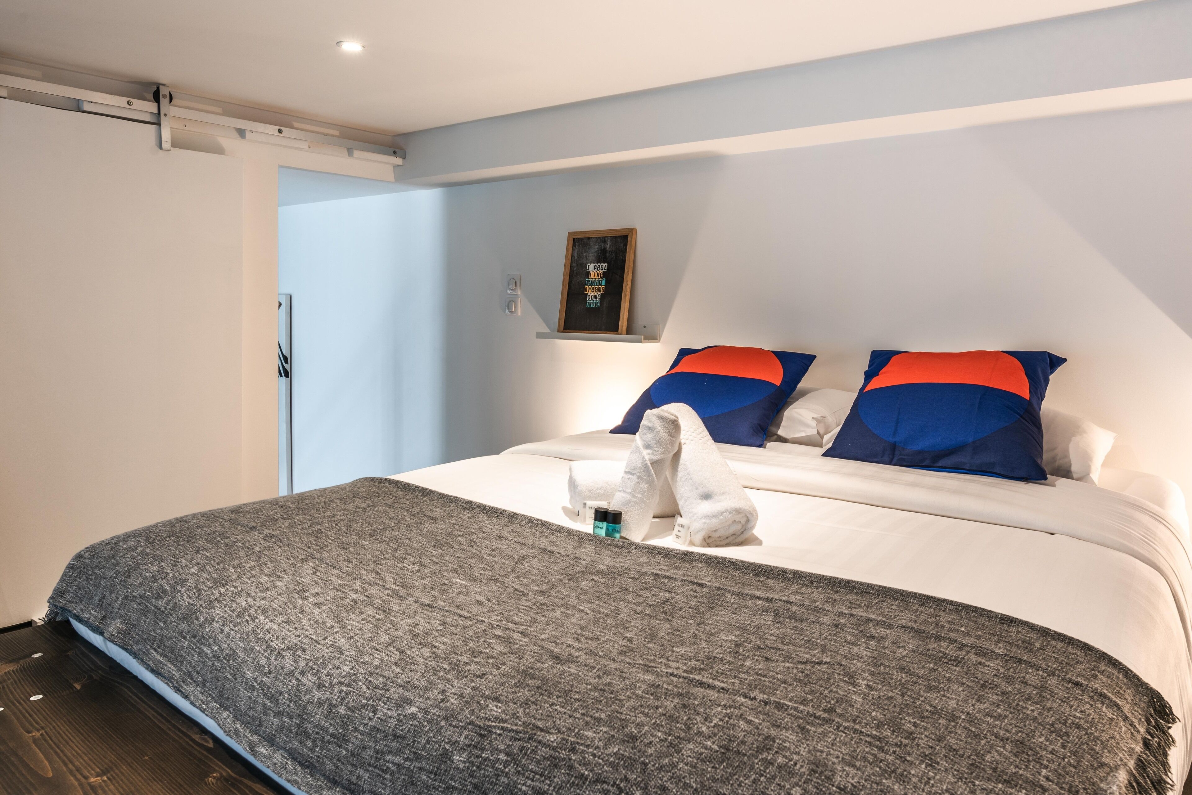 A neatly made bed with a grey blanket, two pillows with blue and red accents, and a small framed item on the wall.