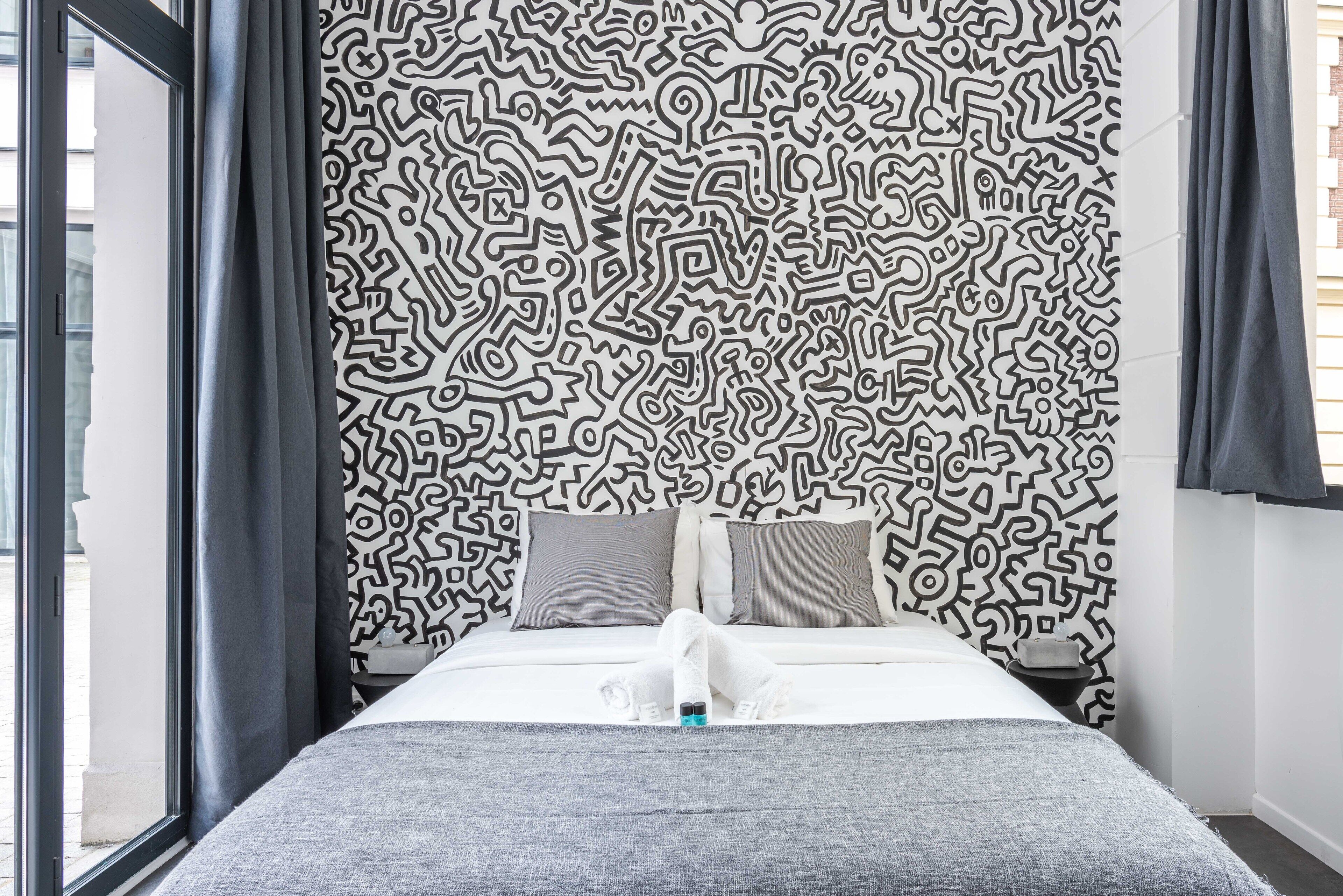 Une chambre d’hôtel avec un papier peint à motifs, un lit avec une literie grise et une fenêtre avec des rideaux.