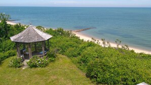 En overdækket pavillon med skråt tag og bænk, med udsigt over en strand med klart blåt vand og en stenet kystlinje.