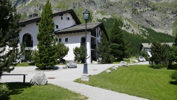 Un bâtiment blanc doté d’un toit en pente, entouré d’une pelouse bien entretenue et avec une chaîne de montagnes en arrière-plan.