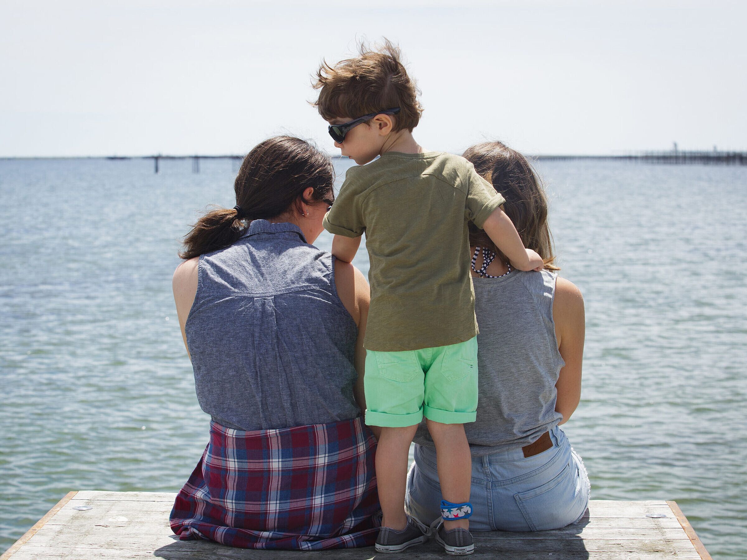 Three people sitting on a wooden dock by a body of water.