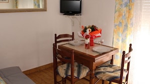 Un coin repas avec une table en bois, quatre chaises, un bouquet de fleurs, une bouteille de vin et un téléviseur fixé au mur.