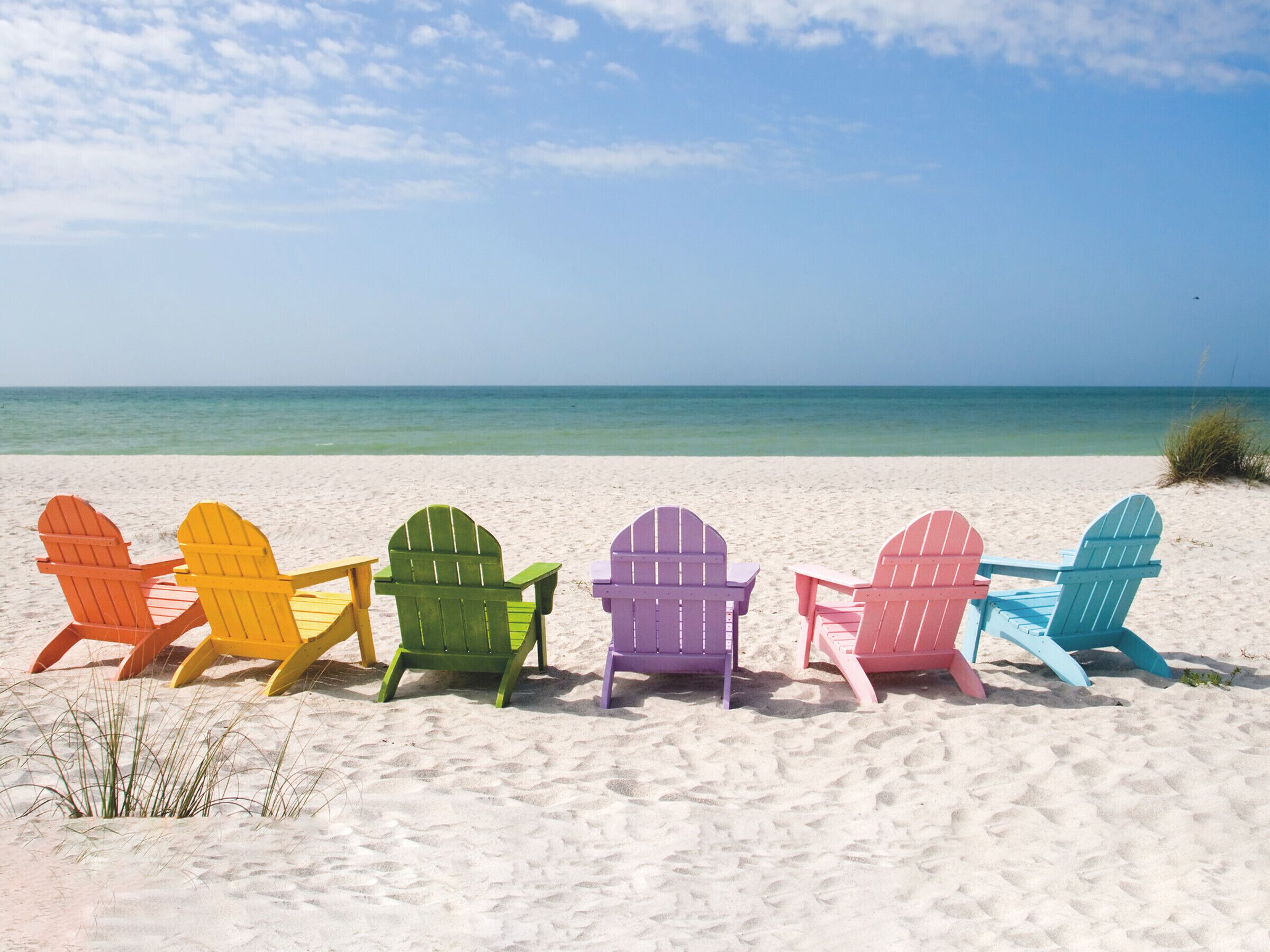 Une rangée de chaises Adirondack aux couleurs vives, sur une plage de sable, avec l’océan en arrière-plan.