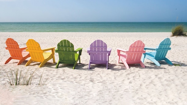 Une rangée de chaises Adirondack aux couleurs vives, sur une plage de sable, avec l’océan en arrière-plan.