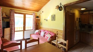 Um quarto acolhedor com teto de madeira, uma mesa de jantar com cadeiras, um sofá com almofadas estampadas, uma janela com cortinas e uma área de cozinha com armários e pia.
