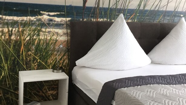 A bedroom with a view of the ocean, featuring a bed with white pillows, a bedside table with a lamp, and a small shelf. - FeWo WeserWeitblick in the 20. Floor in the Columbus Center  (Mitte)