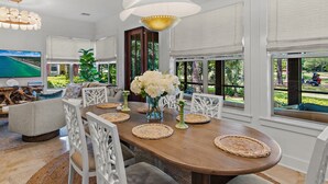 A dining area with a wooden table, white chairs, woven placemats, a vase with white flowers, and a large window with a view of greenery. - Luxury Bayside Sandestin, Beach, 2 Golf Carts (Miramar Beach)