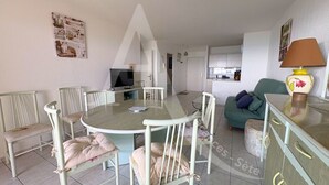 Ein Essbereich mit einem runden Tisch und weißen Stühlen, einer Couch mit Kissen sowie einer Lampe auf einem Schrank.