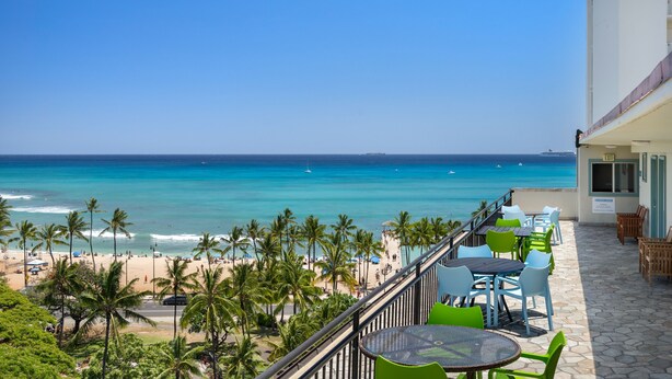 A balcony with a view of the beach and ocean, outdoor seating, and a building entrance.