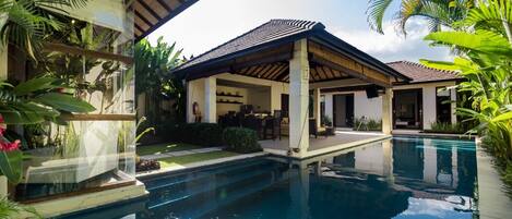 A pool area with a covered patio, a dining area, and lush greenery.