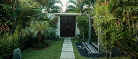 A pathway leading to a dark door surrounded by lush greenery and a small garden area.