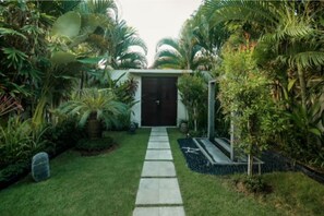A pathway leading to a dark door surrounded by lush greenery and a small garden area.