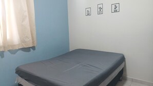 Ein kleines Zimmer mit einem Einzelbett, einer grauen Matratze, einer blauen Wand und einem weißen Vorhang. An der Wand hängen drei gerahmte Bilder.