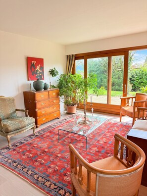 A room with a large window, a wooden cabinet, a green armchair, a patterned rug, and a potted plant. - Homerez - House in Reignier-Esery (Reignier-Esery)