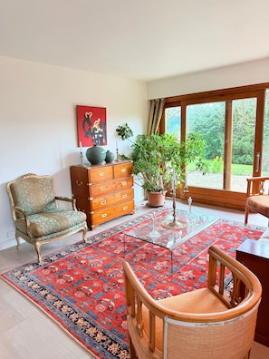 A room with a large window, a red patterned rug, a wooden dresser, a chair, and a potted plant. - Homerez - House in Reignier-Esery (Reignier-Esery)