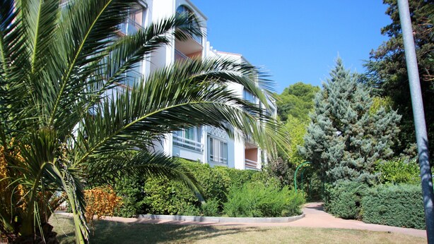 A multi-story building with balconies, surrounded by well-maintained greenery and a clear sky.