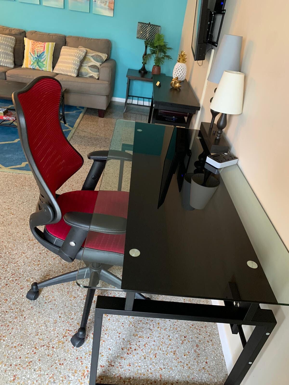 Un espacio de oficina moderno con un escritorio de cristal, una silla de oficina roja, un sofá gris y una mesita con una lámpara.