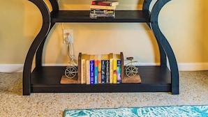 Une console noire sur laquelle sont posés un tableau encadré, une lampe, un ananas et une bibliothèque remplie de livres et surmontée d’un petit vélo.
