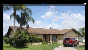 A single-story house with a brown roof, a front porch, and a red car parked in the driveway. - Beautiful View of Intersecting Canals from the Moment You Walk In (Cape Coral)