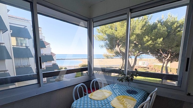 A dining area with a table set for two, large windows offering a view of the sea and a tree-lined path.