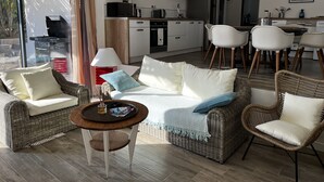 Una sala moderna con muebles de mimbre, un comedor con sillas blancas, una cocina con armarios blancos y un ventanal con cortinas.