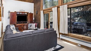 En hyggelig stue med en grå sofa, et træskab og et stort vindue med udsigt over en skov og en grill udenfor.