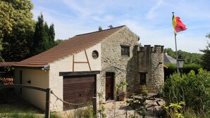 Edifici de pedra amb teulada vermella, un àstal amb una bandera i una zona de jardí amb plantes i una banca.