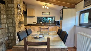 Zona de dinar amb taula de vidre, cadires de fusta, paret de pedra i vistes a la cuina.