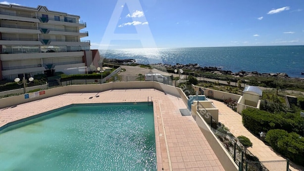 A pool area with a view of the sea, a multi-story building, and a clear sky. - 2 CHAMBRES, LARGE TERRASSE, 1ERE LIGNE MER (Sète)
