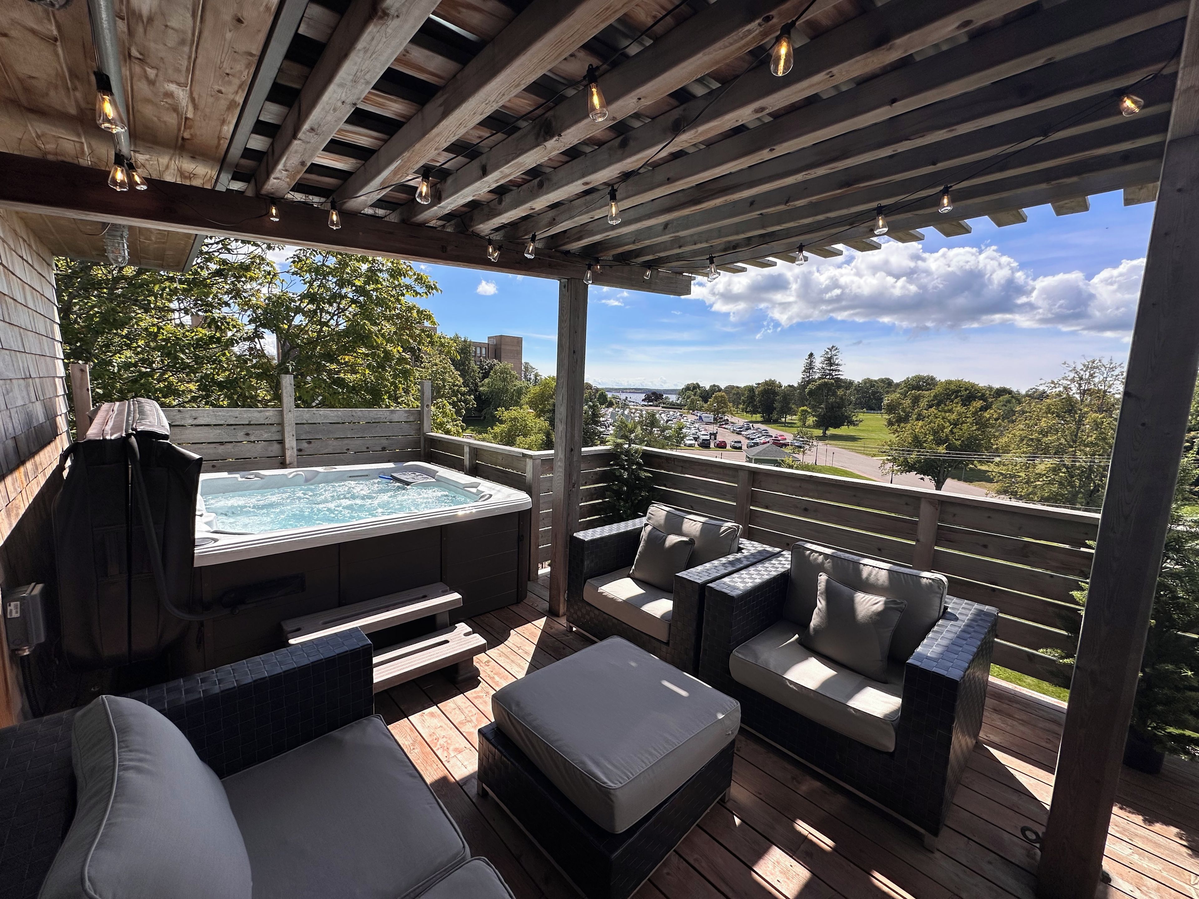Une terrasse en bois avec un jacuzzi, des sièges d’extérieur et une vue sur un parking et des arbres.