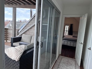 A room with a sliding glass door leading to a balcony, a bed with a headboard, and a small seating area with a cushion and blanket.