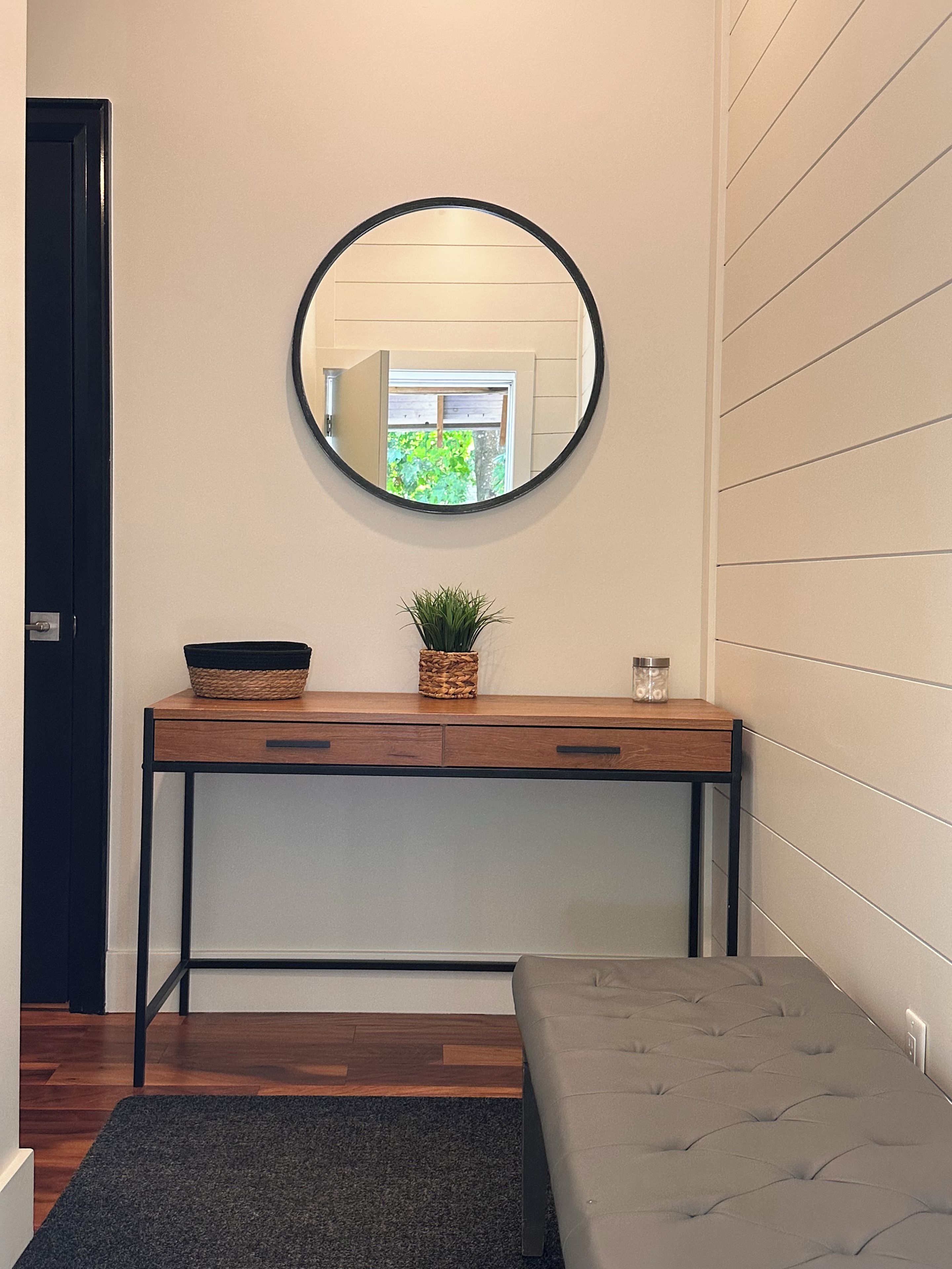A room with a wooden console table, a round mirror, a potted plant, a small container, and a bench.